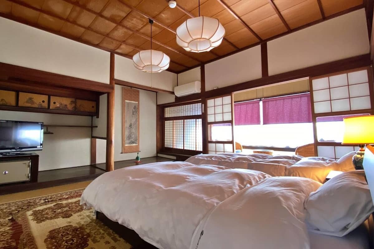 Ein traditioneller japanischer Raum mit Tatami-Bodenbelag, einem Bett mit weißer Bettwäsche, einem Fernseher und einem Fenster mit Blick.