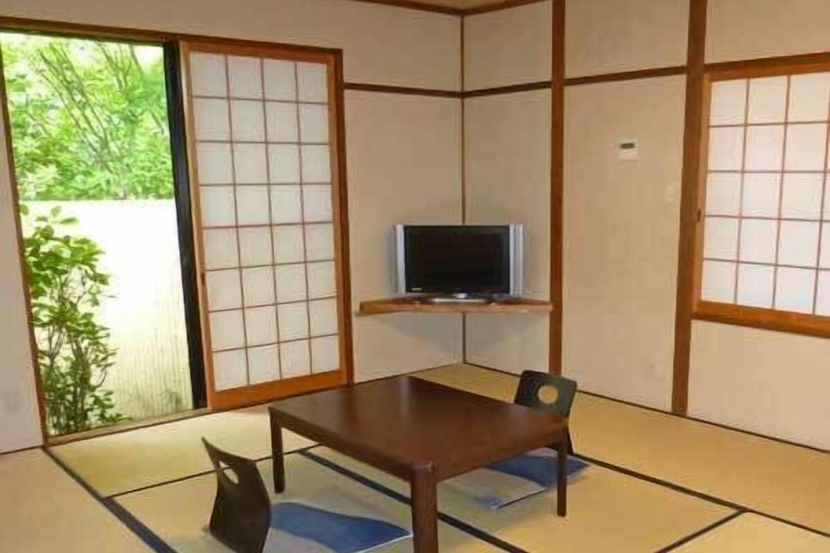 Una habitación con tatami, una mesa baja, dos sillas, un televisor en un estante y una puerta corrediza.