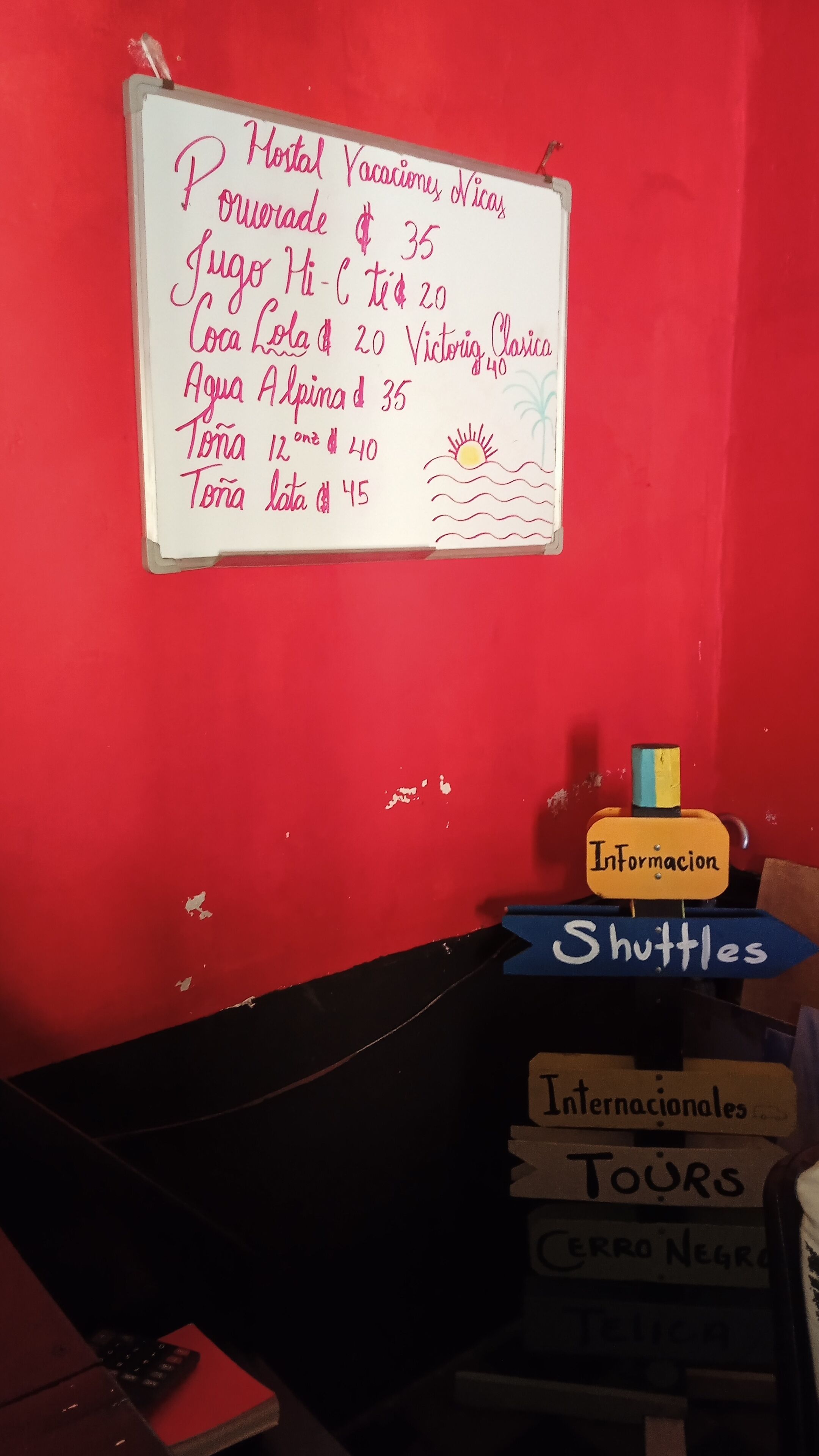 Un tableau blanc affichant une liste d’articles et leurs prix, un petit bureau avec un clavier et un panneau indiquant les services de navette.