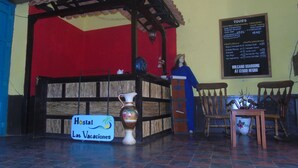 Zona de recepción con un cartel que indica 'Hostal Las Vacaciones', un mostrador de madera, una silla y una pared con una pizarra de menú.