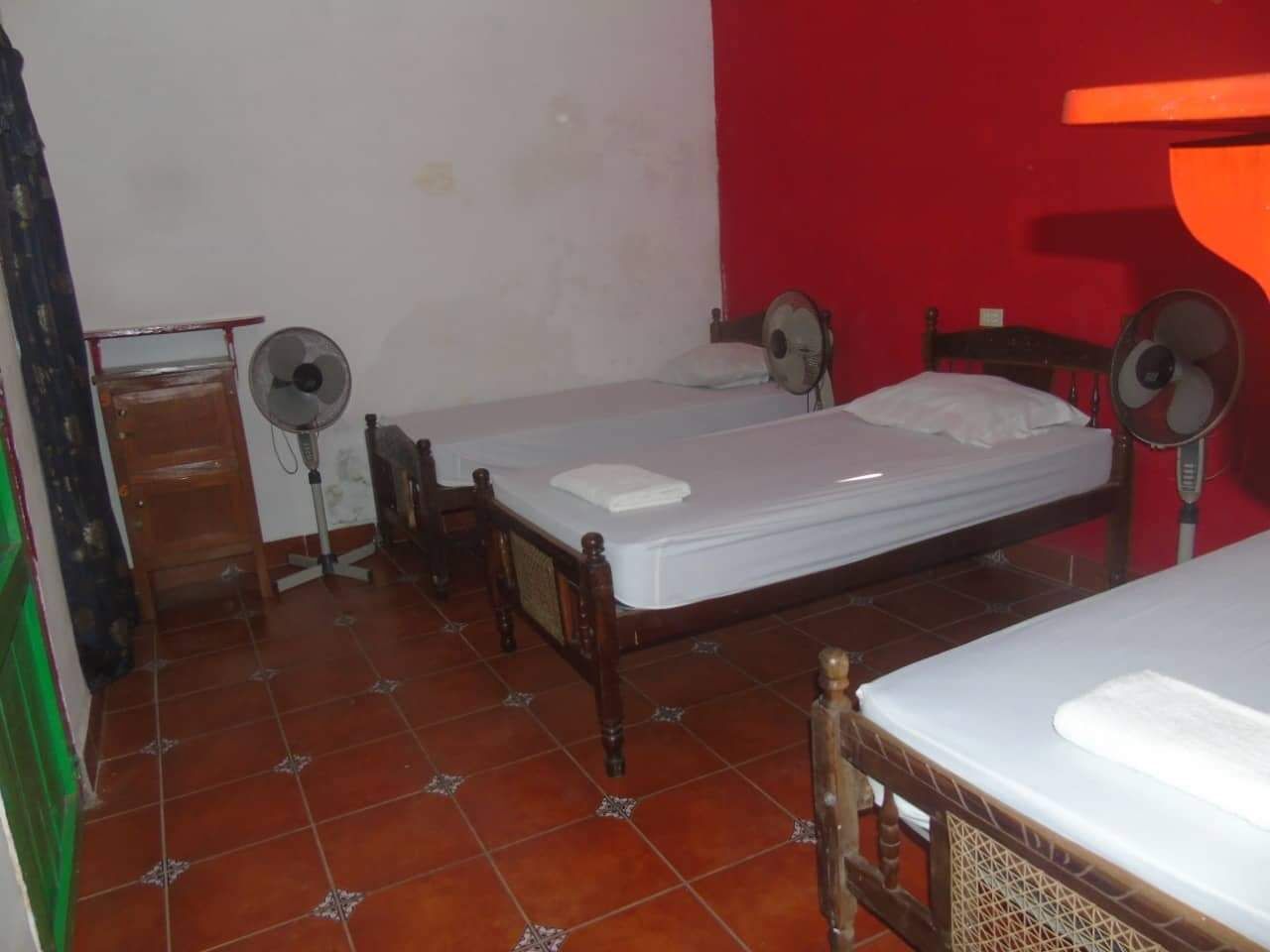 A room with two single beds, a wooden dresser, and two standing fans.