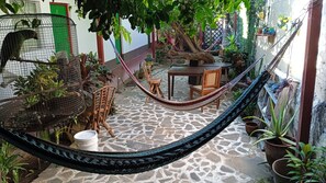Una hamaca en un patio con suelo de baldosas, rodeada de macetas con plantas y una silla de madera.