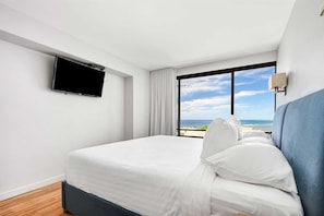 A hotel room with a bed, a TV mounted on the wall, a window with a view of the ocean, and a nightstand with a lamp.
