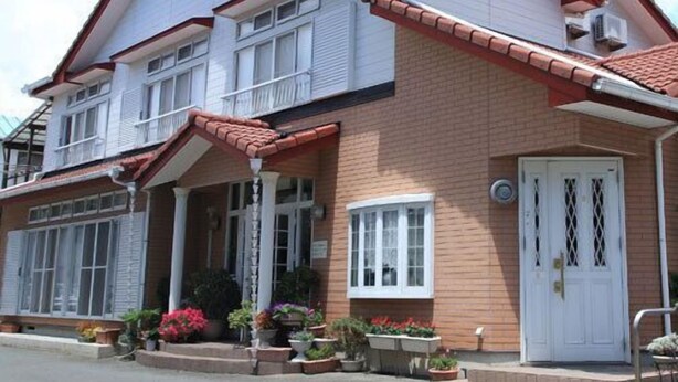 A two-story building with a red-tiled roof, white-framed windows, and a white door with a decorative grille. - Yumigahama Ichibankan (Minamiizu)