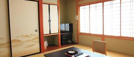 A traditional Japanese room with tatami flooring, a low table with a cushion, a television, and sliding doors.