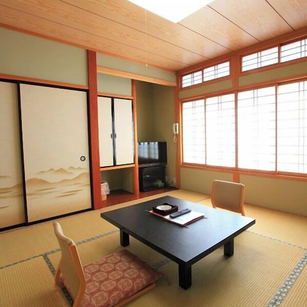Une pièce de style japonais traditionnel, avec un sol recouvert de tatamis, une petite table basse avec un coussin, un téléviseur et des portes coulissantes.