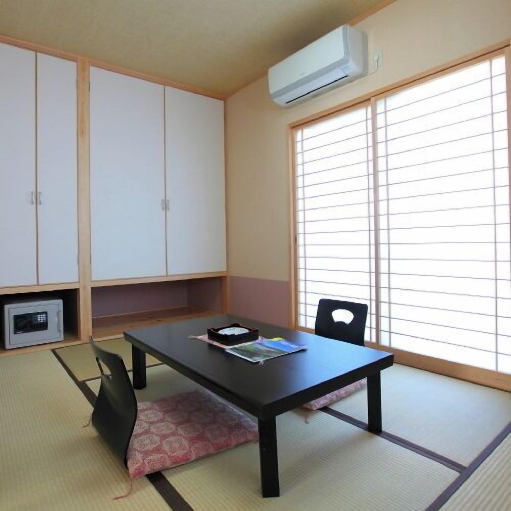 A room with tatami flooring, a black table with chairs, a window with blinds, and an air conditioning unit.