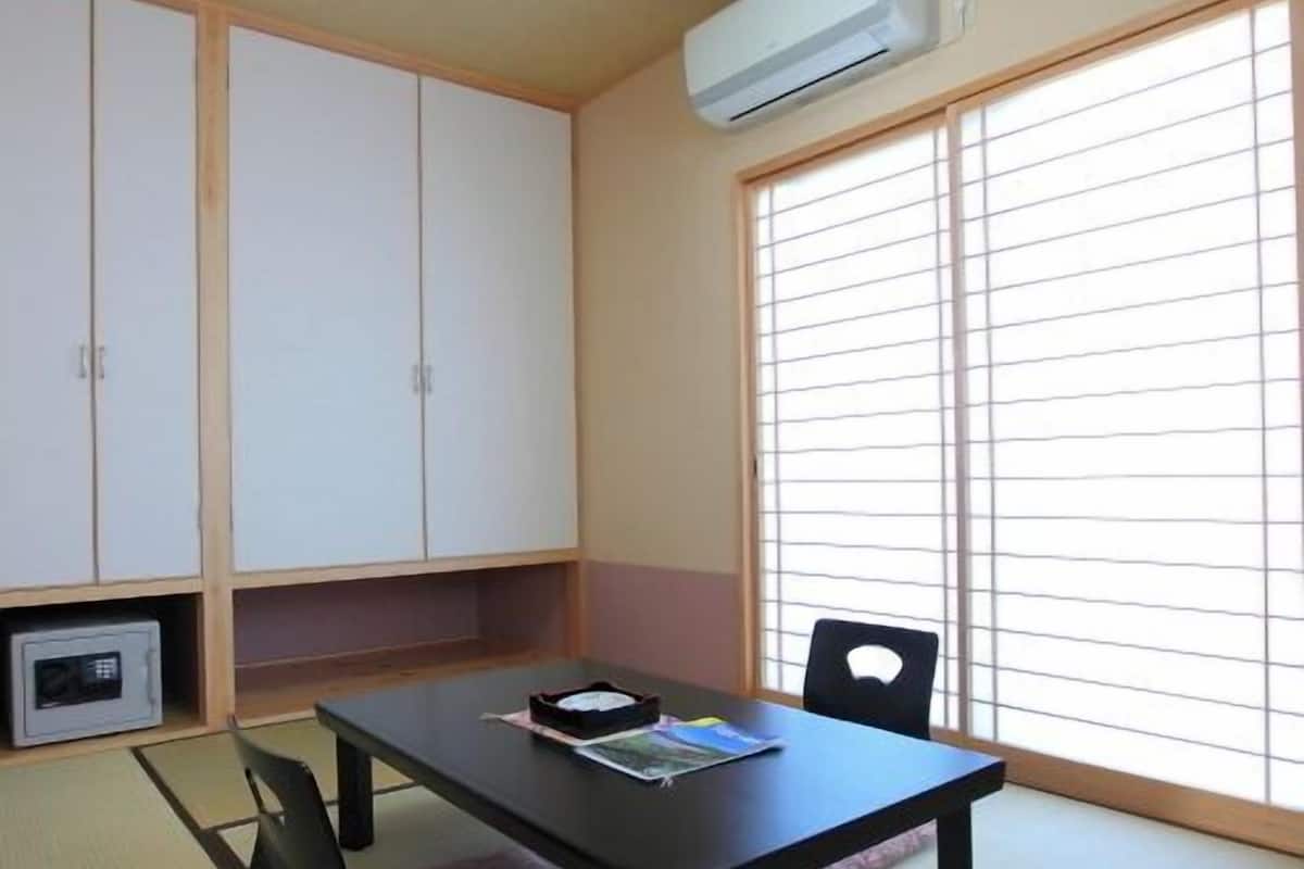 A room with tatami flooring, a black table with chairs, a window with blinds, and an air conditioning unit.