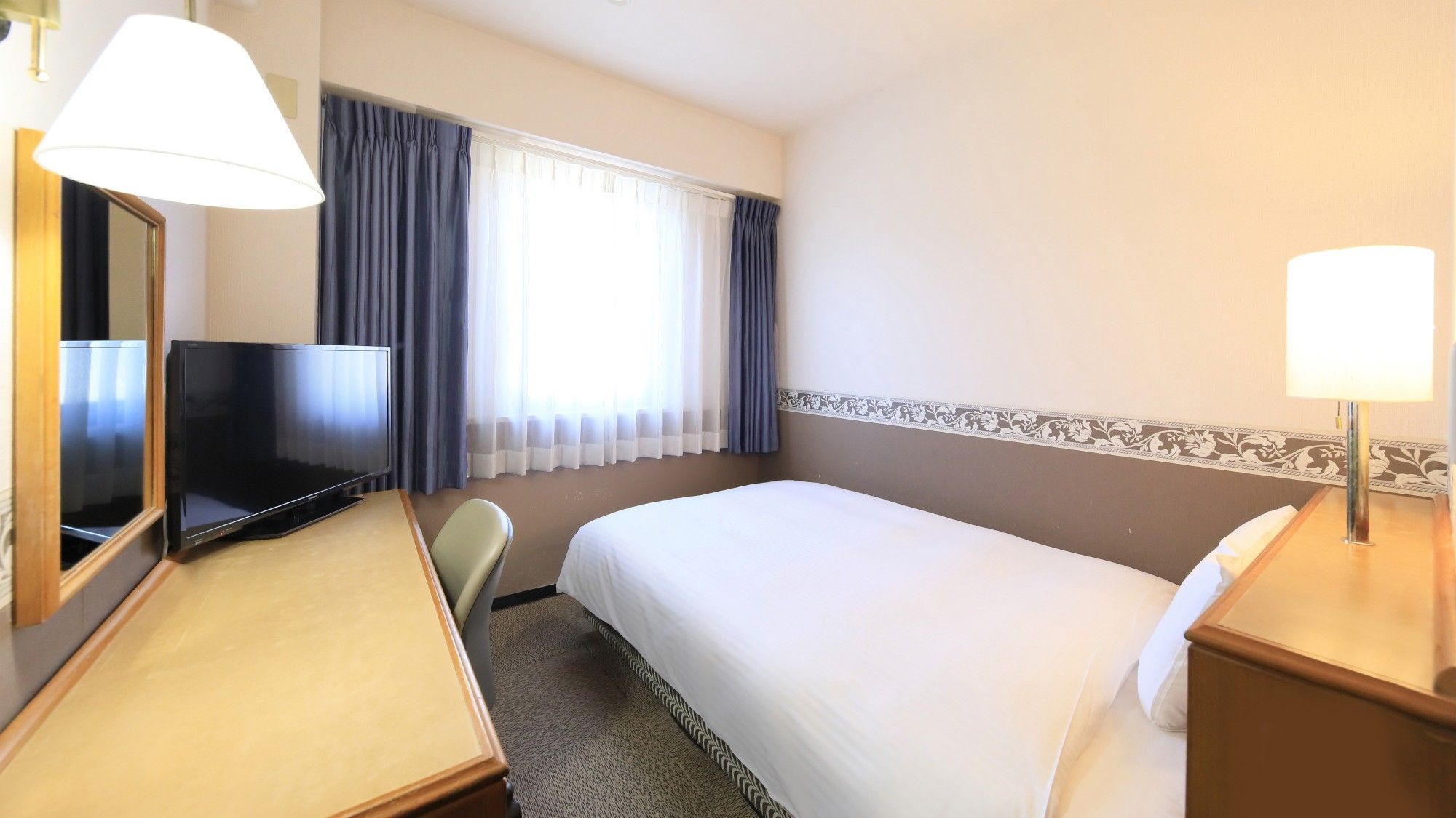A hotel room with a bed, a desk, a TV, a lamp, and a window with curtains.