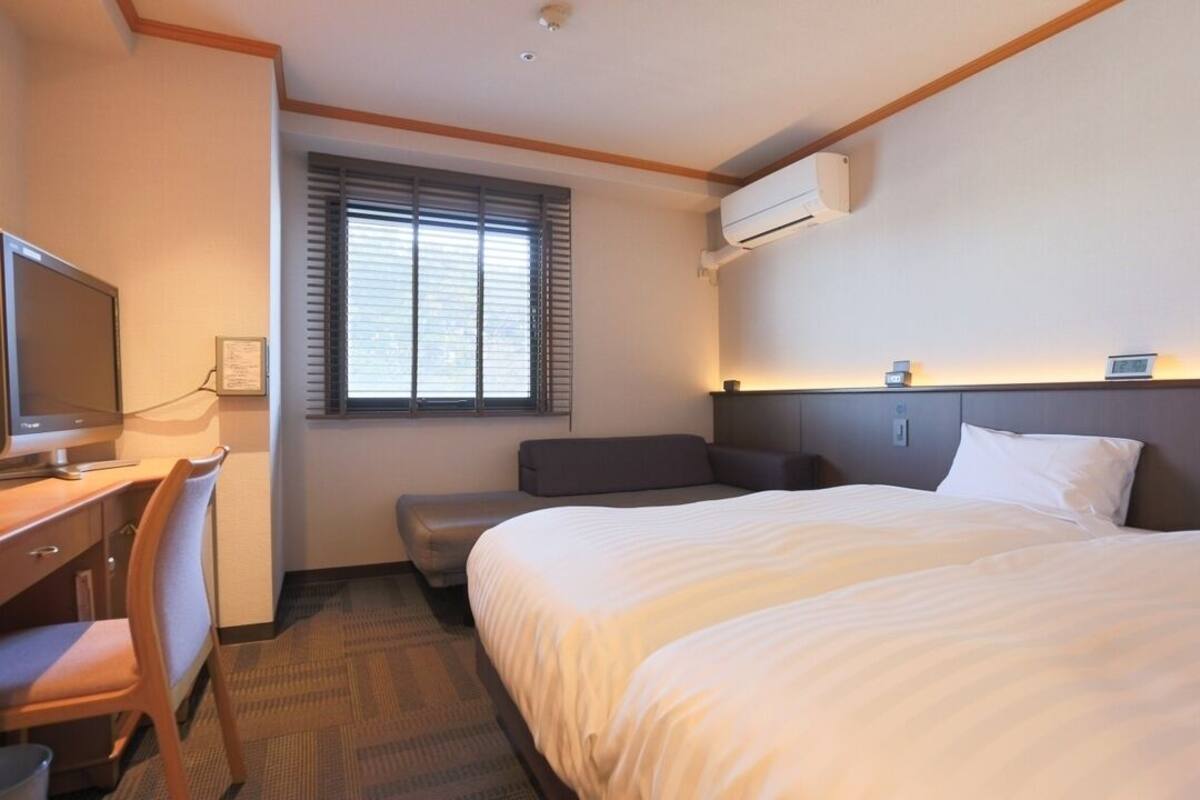 A hotel room with a bed, a desk with a chair, a television, and a window with blinds.