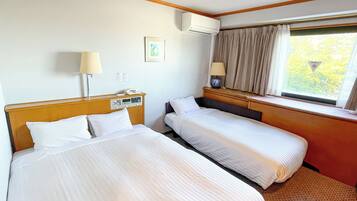 A hotel room with two beds, a bedside table with a lamp, a window with curtains, and an air conditioning unit on the wall.