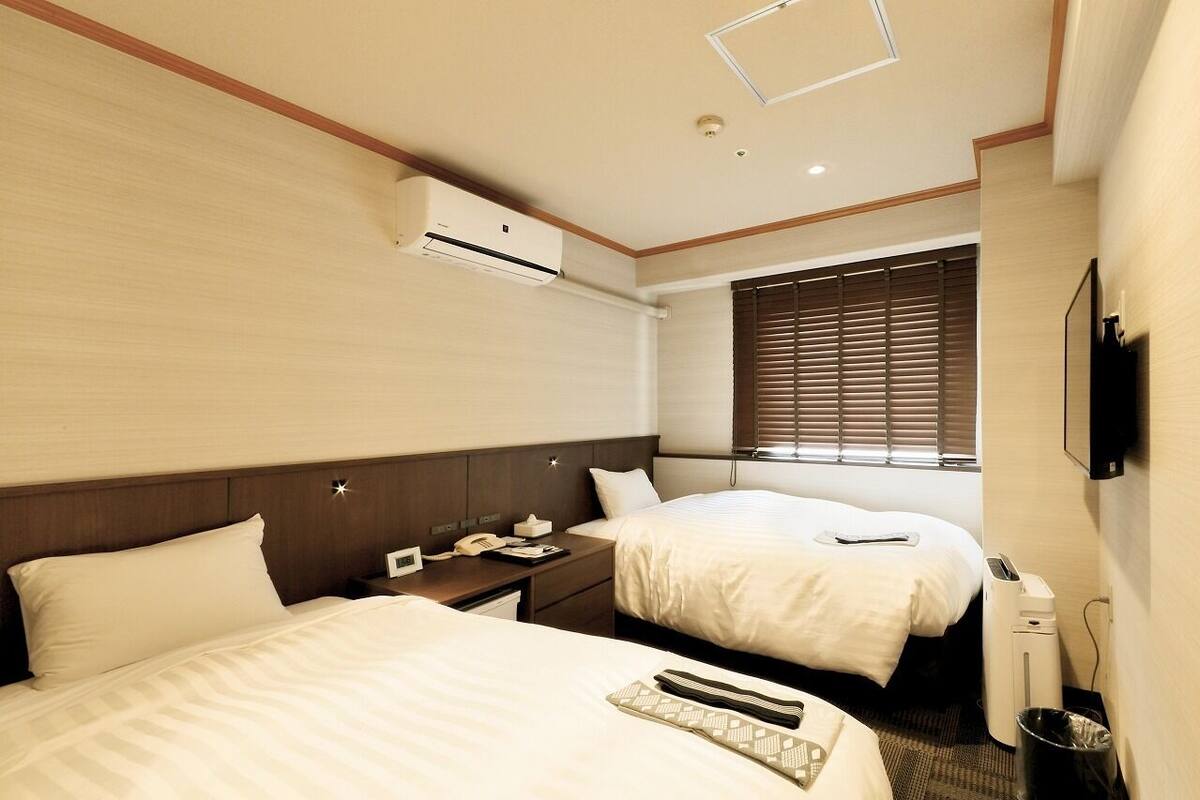 A hotel room with two beds, a desk, an air conditioning unit, a TV, and a window with blinds.