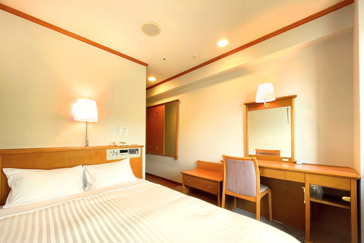 A hotel room with a bed, a desk, a chair, a mirror, and a lamp.