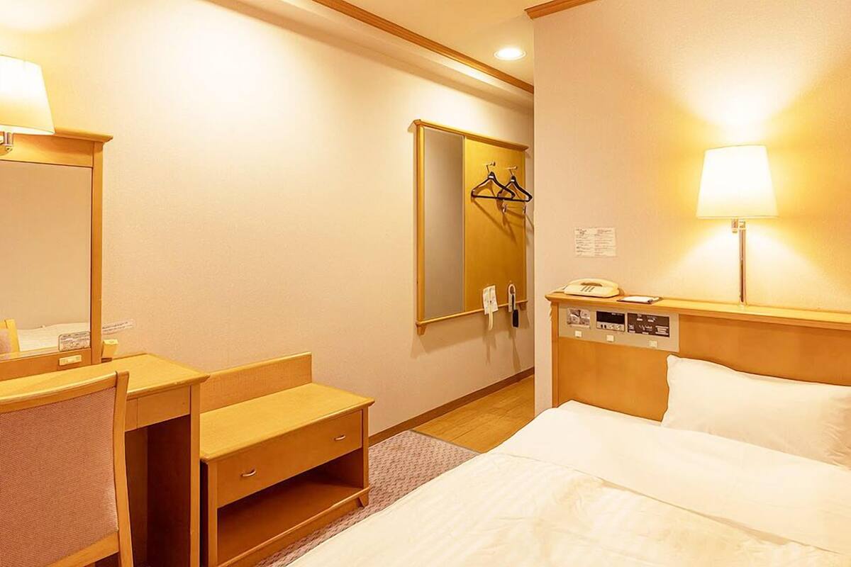 A hotel room with a bed, a desk, a chair, a mirror, a wardrobe, and a bedside table with a lamp.