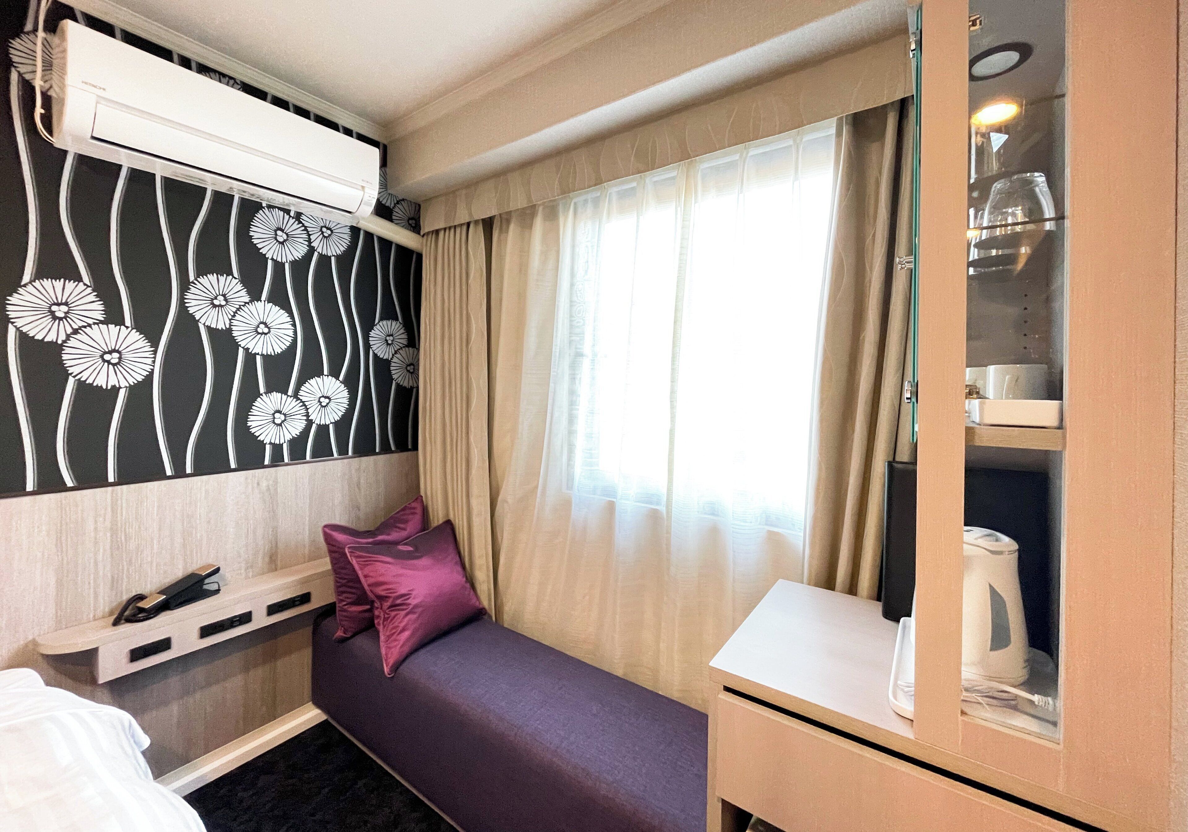 A compact hotel room with a bed, a small desk, a built-in air conditioner, a window with curtains, and a minibar.
