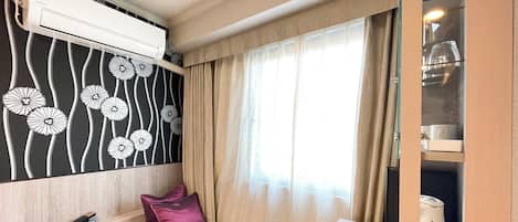 Habitación de hotel pequeña con cama, escritorio, aire acondicionado integrado, ventana con cortinas y minibar.