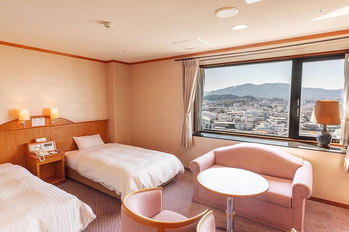 A hotel room with two beds, a small round table, a chair, a nightstand with a phone, and a window with a city view.