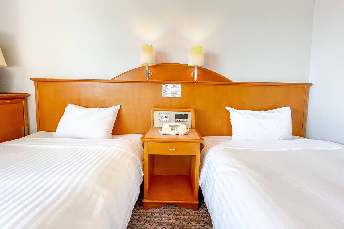 A hotel room with two single beds, a wooden headboard, bedside tables with lamps, a clock, and a telephone.