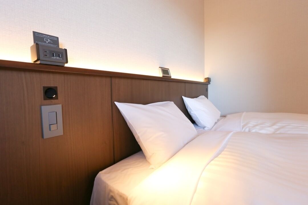 A hotel room with a wooden headboard, a bedside table with a clock and a power outlet, and a bed with white bedding.
