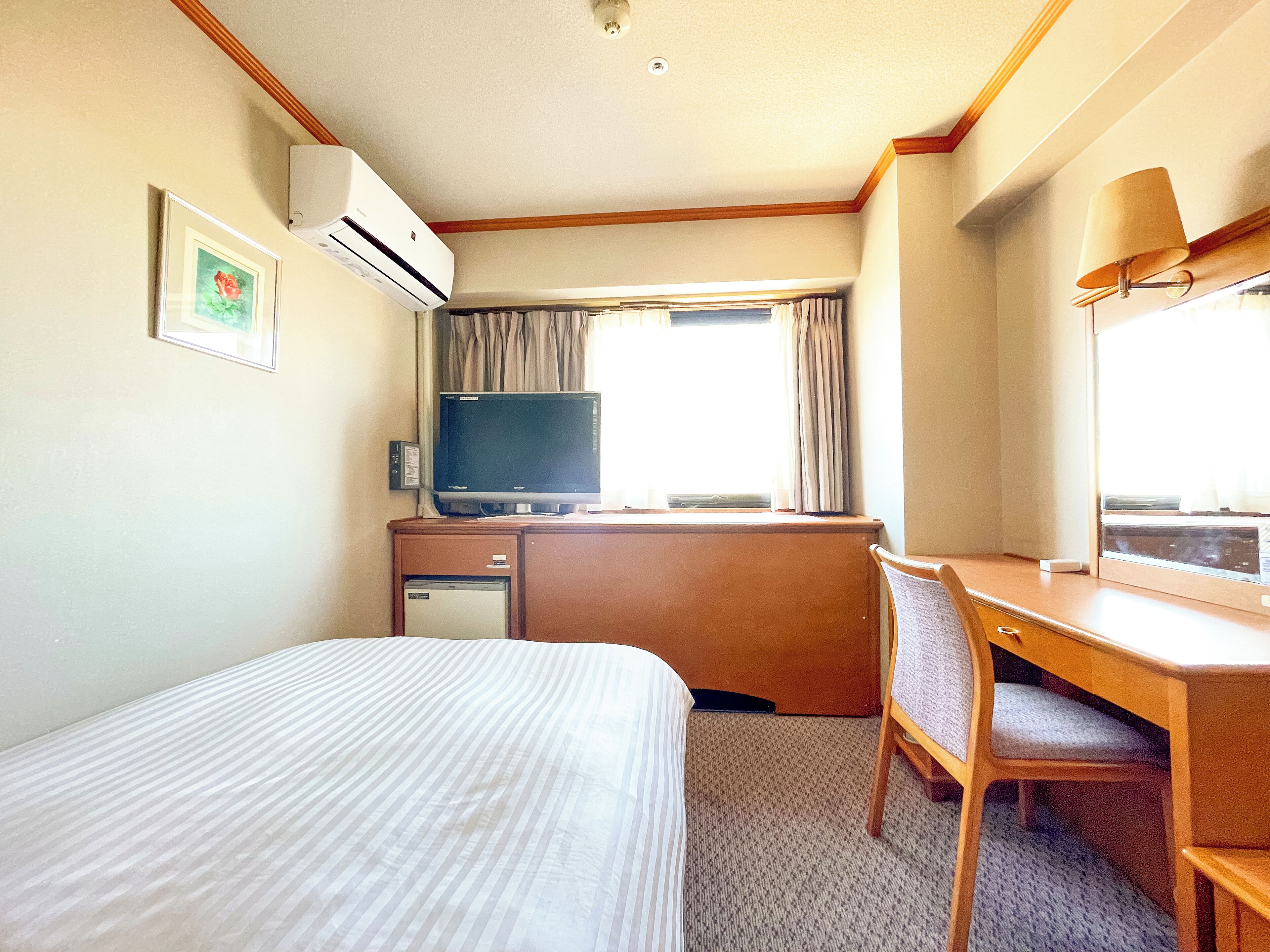 A hotel room with a bed, a desk, a chair, a television, an air conditioner, and a window with curtains.