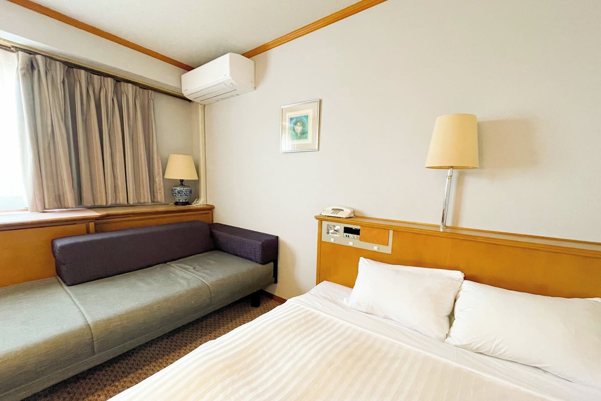 A hotel room with a bed, a sofa, a bedside table with a lamp, a window with curtains, and an air conditioning unit.