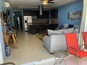 Ein modernes Wohnzimmer mit einem grauen Sofa, roten Sesseln und einem Glastisch. Es gibt einen Küchenbereich mit dunklen Schränken und einem Edelstahlkühlschrank.