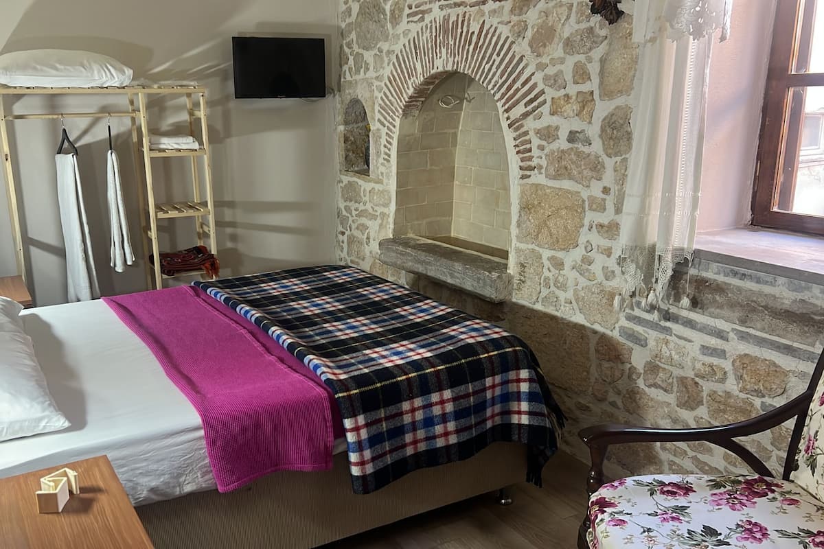 Une pièce avec un lit, une étagère en bois où sont posées des serviettes, un mur en pierre et une fenêtre.