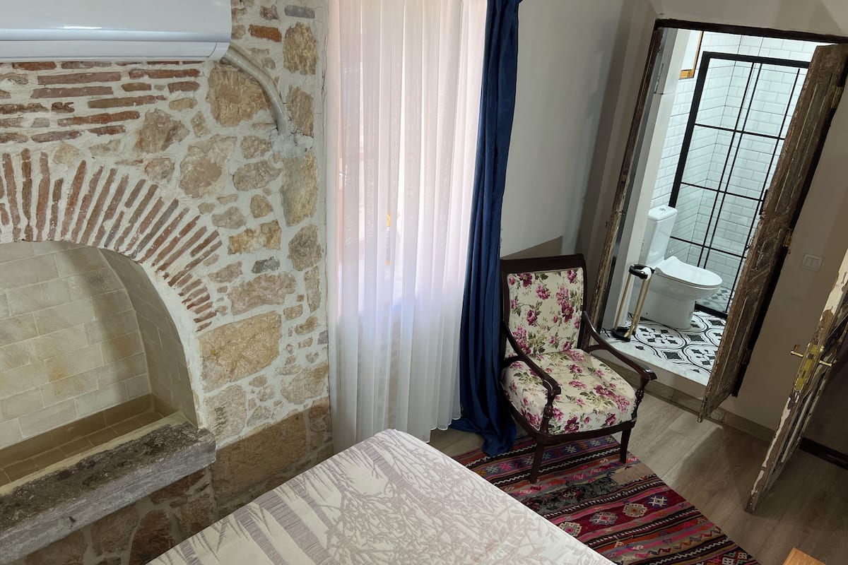 Une pièce avec une cheminée en pierre, un fauteuil à motifs floraux, un tapis à dessins et une vue sur une salle de bain carrelée, visible à travers une fenêtre.