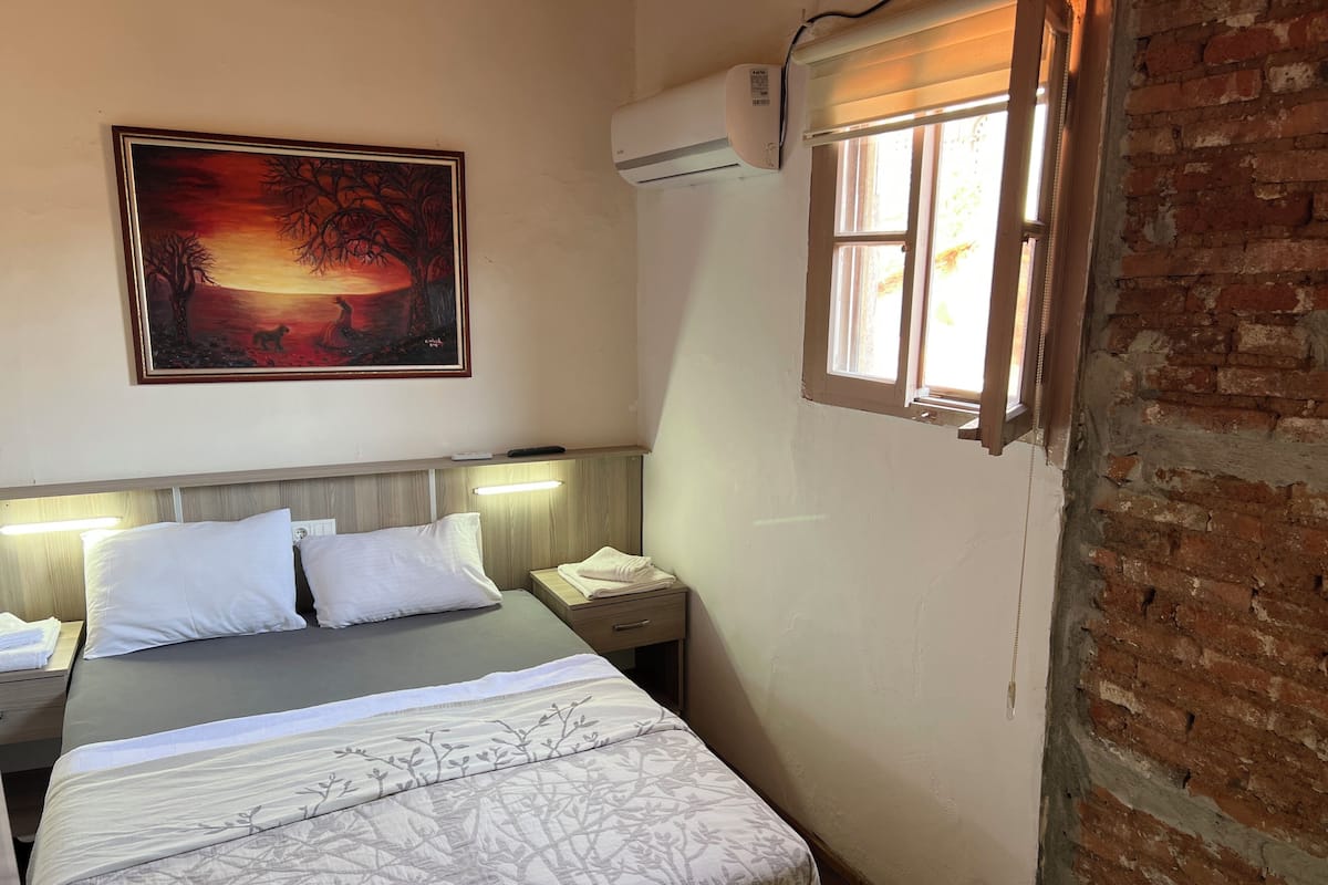 Une chambre d’hôtel comprenant un lit, des tables de chevet, une petite table, un tableau, un appareil de climatisation et une fenêtre donnant sur un mur en briques.