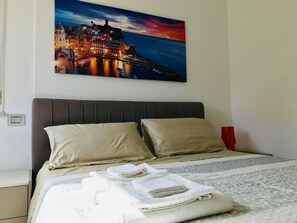 A neatly made bed with beige pillows and white towels, a dark headboard, and a painting of a coastal town at sunset above it. - B&B La Maggiolina (La Spezia)