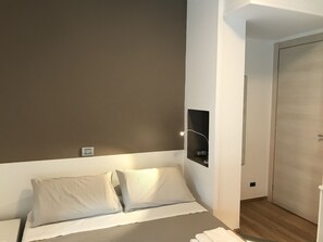 Un dormitorio con cama, dos almohadas, una mesita de noche, una lámpara de pared y una puerta.