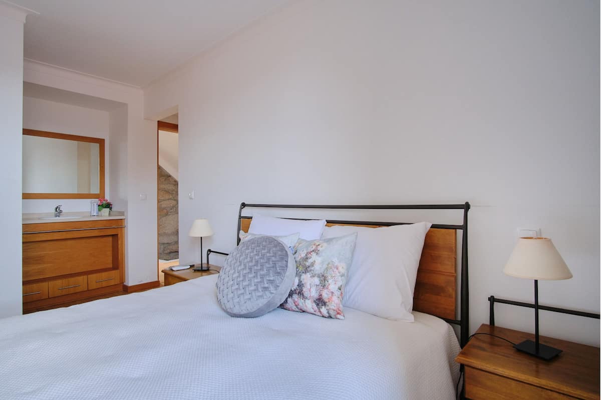 Een net opgemaakt bed met een houten hoofdbord, een nachtkastje met een lamp, en een houten dressoir met een spiegel.