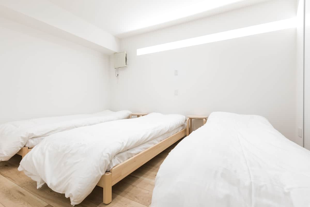 Une pièce avec trois lits en bois alignés contre un mur blanc, une petite table de chevet et un appareil de climatisation fixé au plafond.