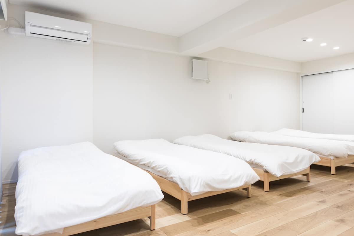 Une pièce avec trois lits en bois alignés contre un mur blanc, un appareil de climatisation blanc fixé au-dessus des lits, et une porte blanche sur le côté droit.