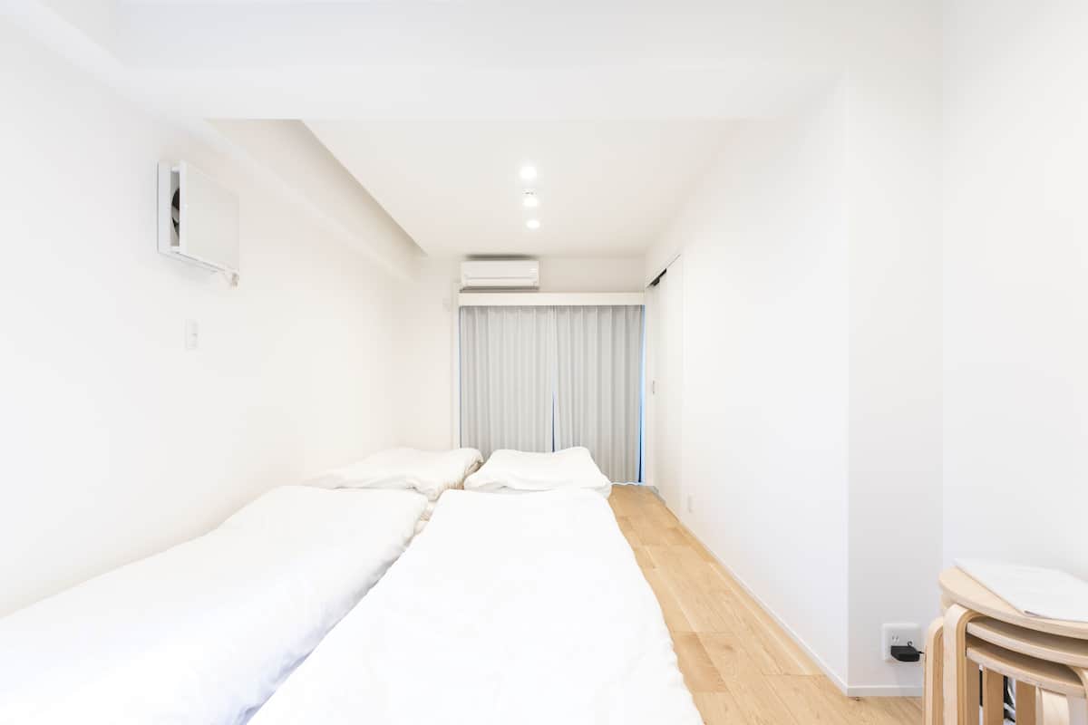 Une chambre d’hôtel minimaliste avec deux lits simples, une petite table en bois, une fenêtre avec des rideaux et un appareil de climatisation.