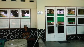 A person sitting on a bench outside a building with a tiled wall and a door with multiple windows. - Olivia Homestay (Banyuwangi)