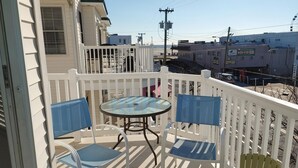 Un balcon avec une table en verre et deux chaises, une clôture blanche et une vue sur un parking et des bâtiments en arrière-plan.