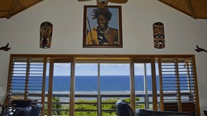 A room with a large window overlooking the ocean, a ceiling fan, and two wooden carvings on the walls.