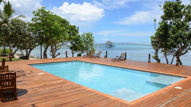 A wooden deck with a swimming pool, lounge chairs, and a view of the ocean and pier.