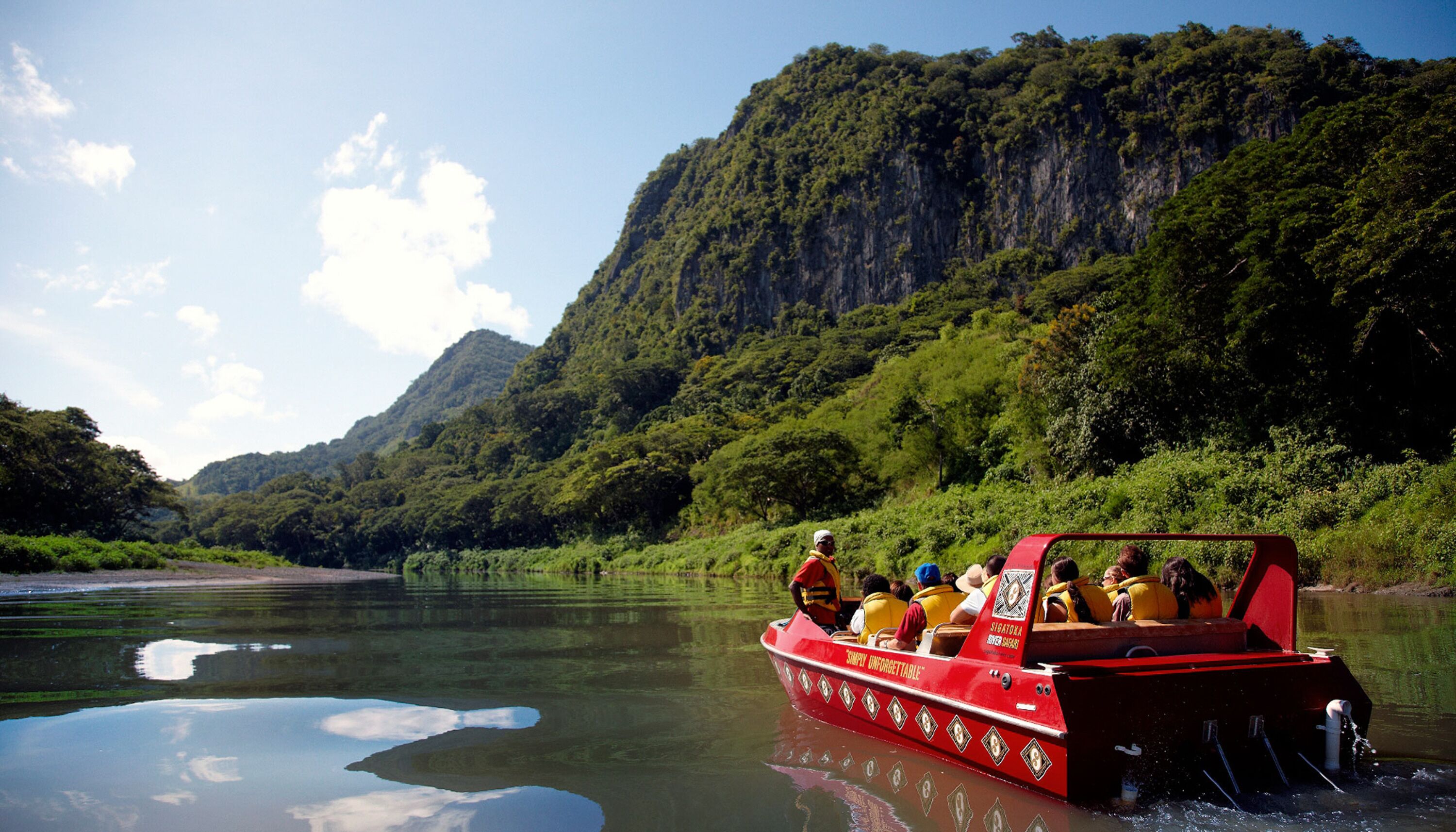A red boat with passengers on a river surrounded by lush greenery and mountains.