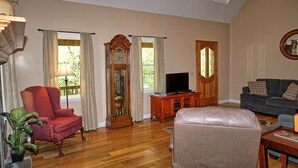 A living room with a red armchair, a large wooden clock, a television, and a grey sofa. - Willow Tree Lodge-Easy access - Secluded - Grill (Bryson City)