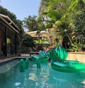 Un espace piscine agrémenté de bouées gonflables en forme de cactus, entouré d’une végétation luxuriante et doté d’un espace salon abrité.