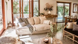 A spacious living room with a beige sofa, a wooden coffee table, and a view of a garden through large sliding glass doors. - Beach Jungle: Pet-Friendly Hot Tub, Firepit & Pool in Beachside Hideaway (Byron Bay)