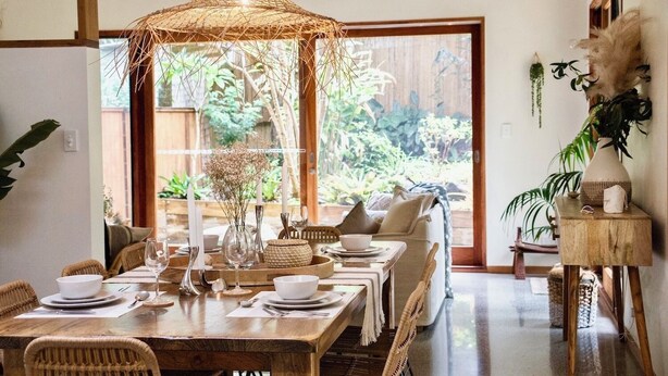 A dining area with a wooden table set for four, surrounded by wicker chairs. A woven ceiling light fixture hangs above, and a large window offers a view of greenery outside. - Beach Jungle: Pet-Friendly Hot Tub, Firepit & Pool in Beachside Hideaway (Byron Bay)