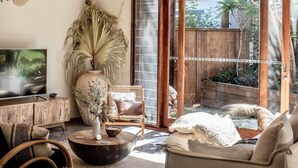 A spacious living room with a large TV, a wooden ceiling, and a sliding glass door leading to a garden. - Beach Jungle: Pet-Friendly Hot Tub, Firepit & Pool in Beachside Hideaway (Byron Bay)
