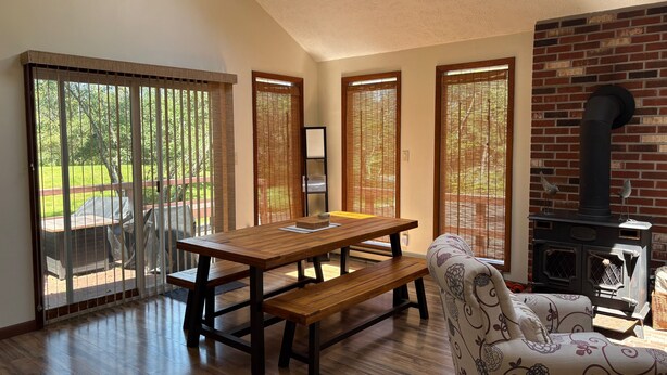 A dining area with a wooden table and benches, a patterned armchair, and a brick fireplace. - Ash Cottage in Poconos near Camelback & Kalahari (Long Pond)