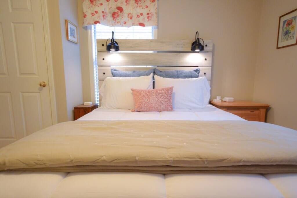 Quarto com cama grande, cabeceira de madeira, duas luminárias laterais e janela com cortinas florais.