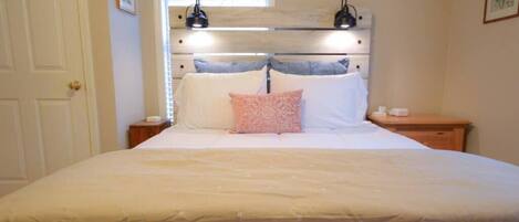 Quarto com cama grande, cabeceira de madeira, duas luminárias laterais e janela com cortinas florais.
