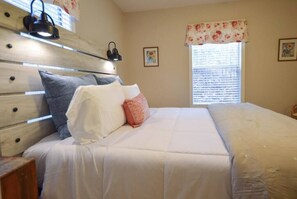 Une chambre à coucher avec une tête de lit en bois, un lit recouvert d’une literie blanche, une fenêtre avec des stores et une lampe fixée au mur.