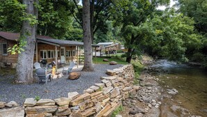 A rustic cabin with a stone wall and a fire pit, surrounded by trees and a river. - Deep Creek Cottage-Creek side- close to the Polar Express (Bryson City)